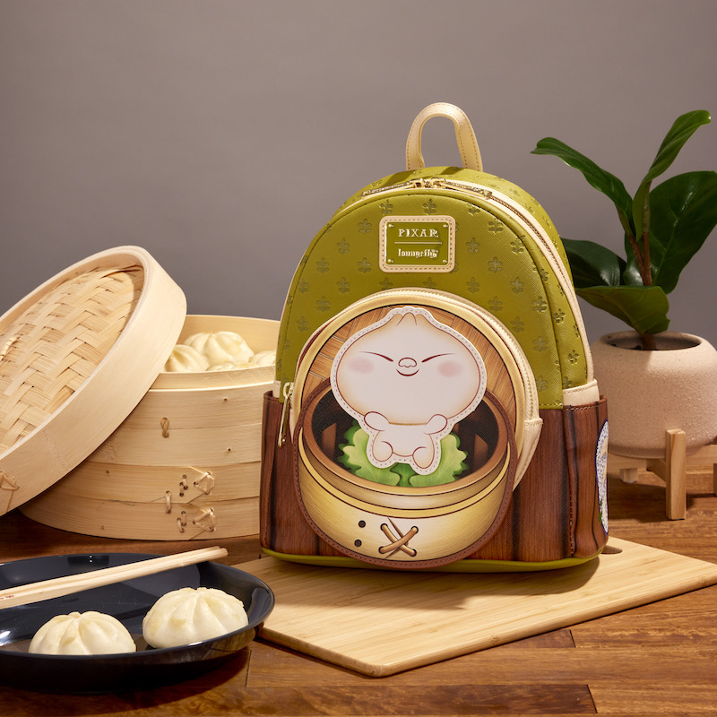 Green and brown Loungefly mini backpack that has a front pocket with a happy Bao on the front sitting on a wooden table surrounded by cooked bao and steamer baskets 